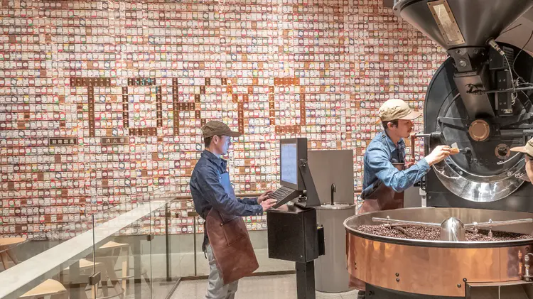 Display of hundreds of colorful Starbucks Reserve coffee cards on a large wall, with "TOKYO" spelled out within, and coffee roasting equipment in foreground